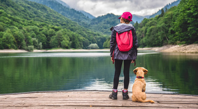 Girl with a small yellow dog standing on pier by the mountain lake